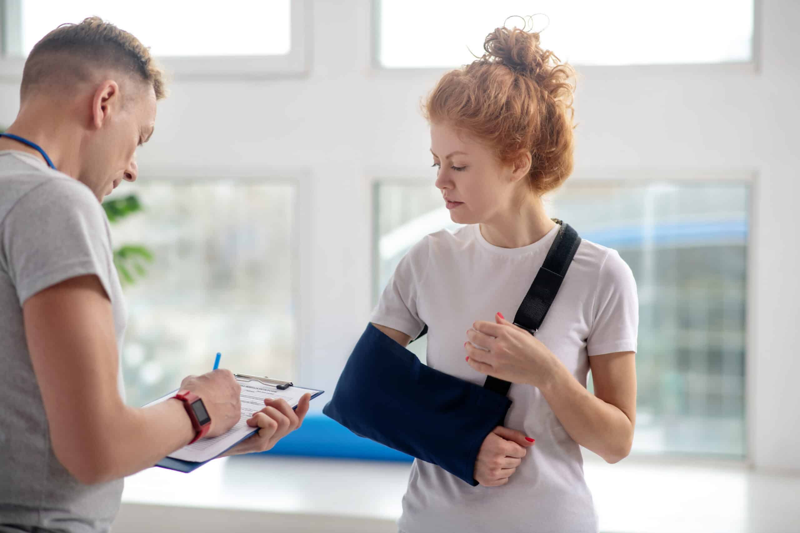 Male physiotherapist making notes, female patient standing in front of him in arm sling